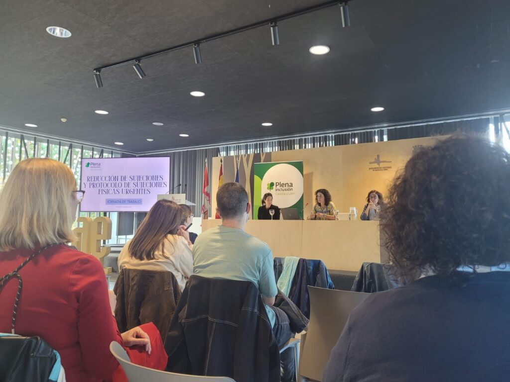 Sesión de trabajo sobre reducción de restricciones. En una mesa presidencial hay tres profesionales de plena inclusión, mientras que se ve varias personas del público a la espalda.